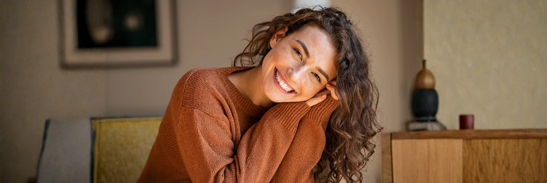 Közép-európai fiatal nő mosolyog a kamerába, barna pulcsit viselve, otthoni környezetben.